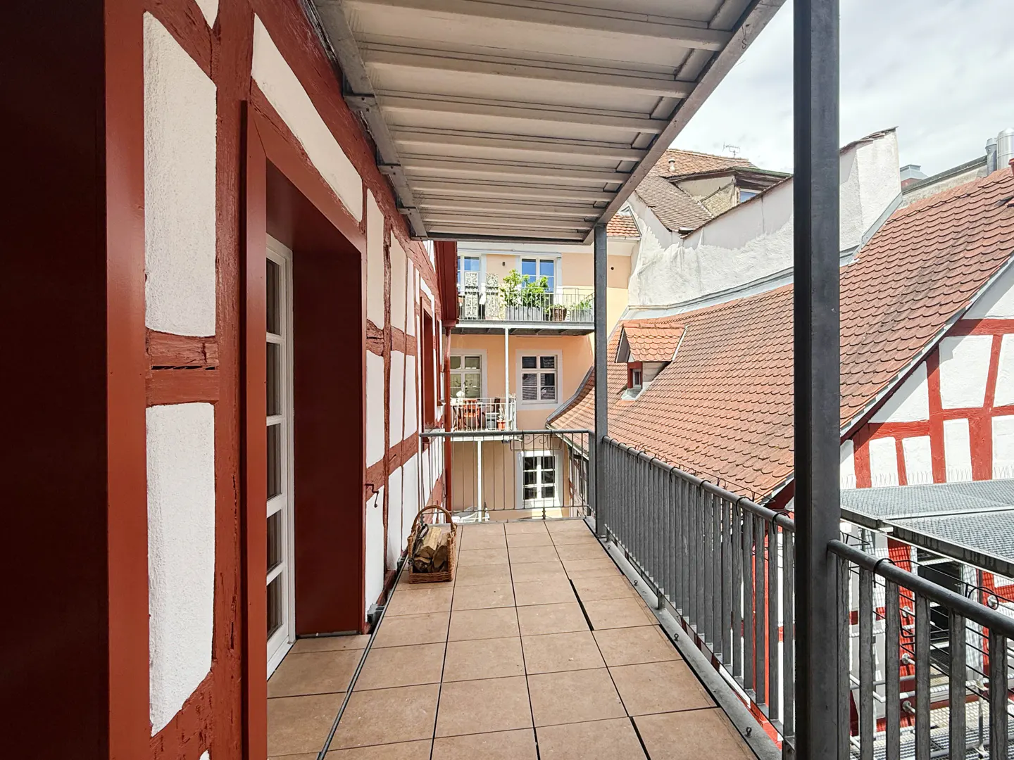 Covered balcony with tiled floor and wood basket, overlooking a courtyard with traditional half-timbered buildings.
