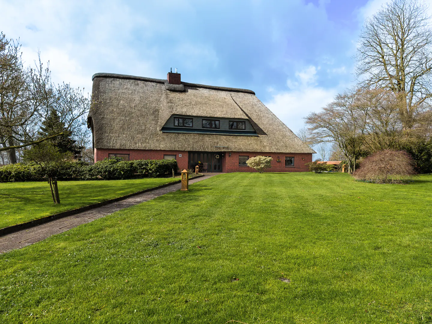 A red brick house with a thatched roof sits on a large green lawn under a blue sky.