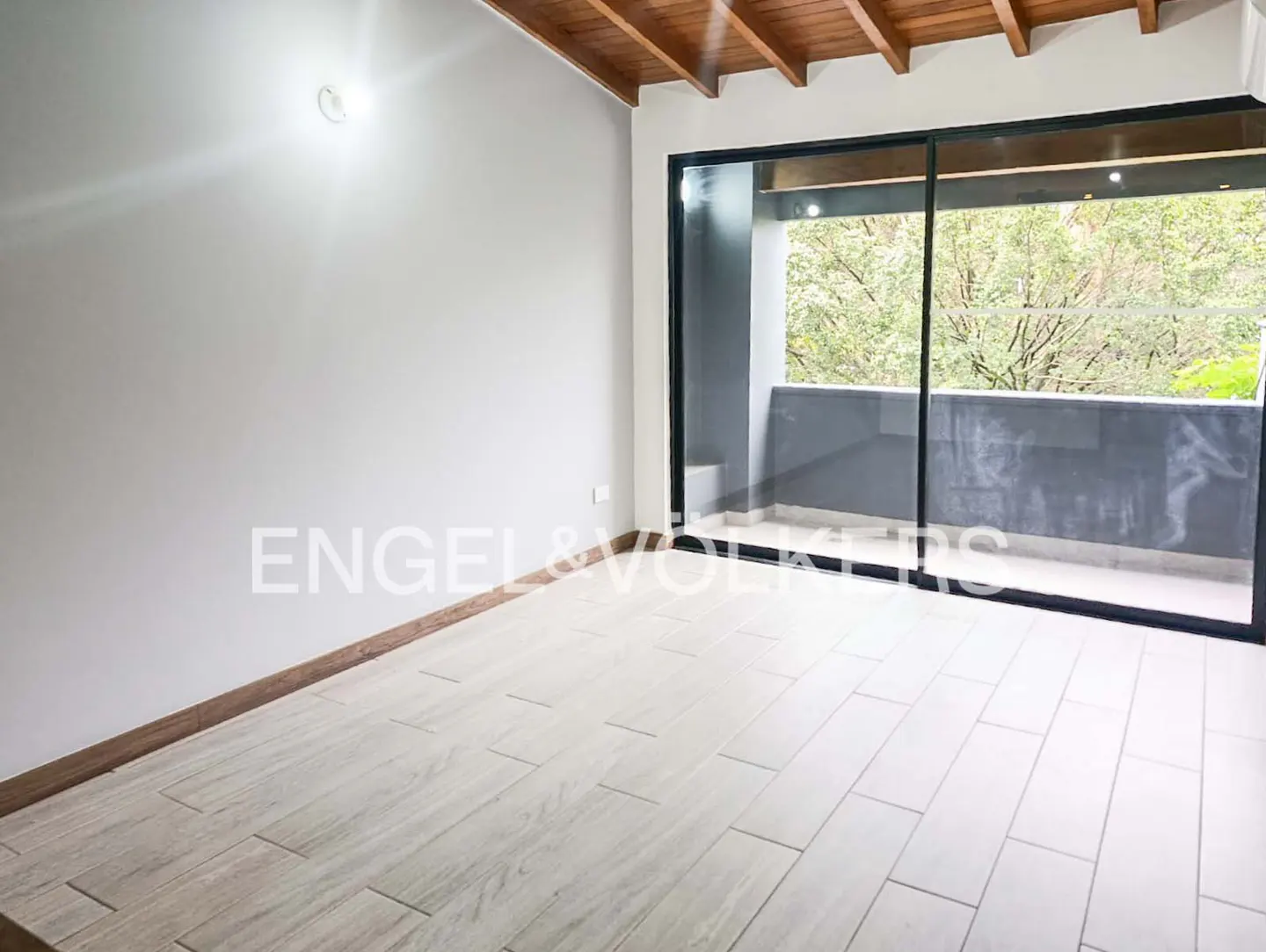 A bright, empty room with white walls, light tile floors, and a large sliding glass door to a balcony with trees visible.