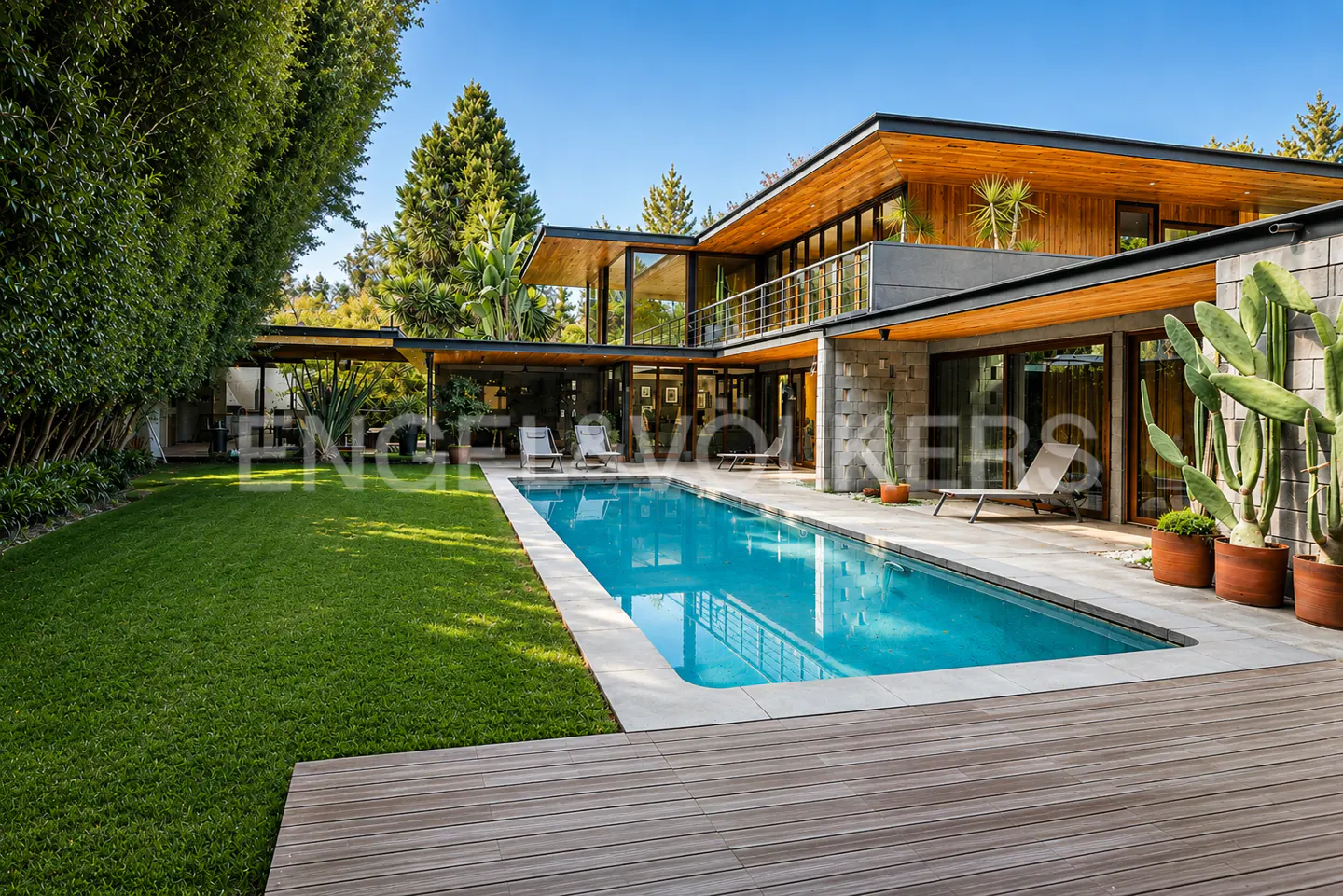 Modern two-story home with a pool. The house has wood and stone accents, large windows, and a balcony. A green lawn and trees surround the property.