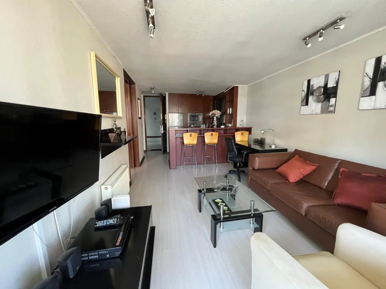 A wide shot of a living room with a brown leather sofa, glass coffee table, and kitchen bar with yellow stools.
