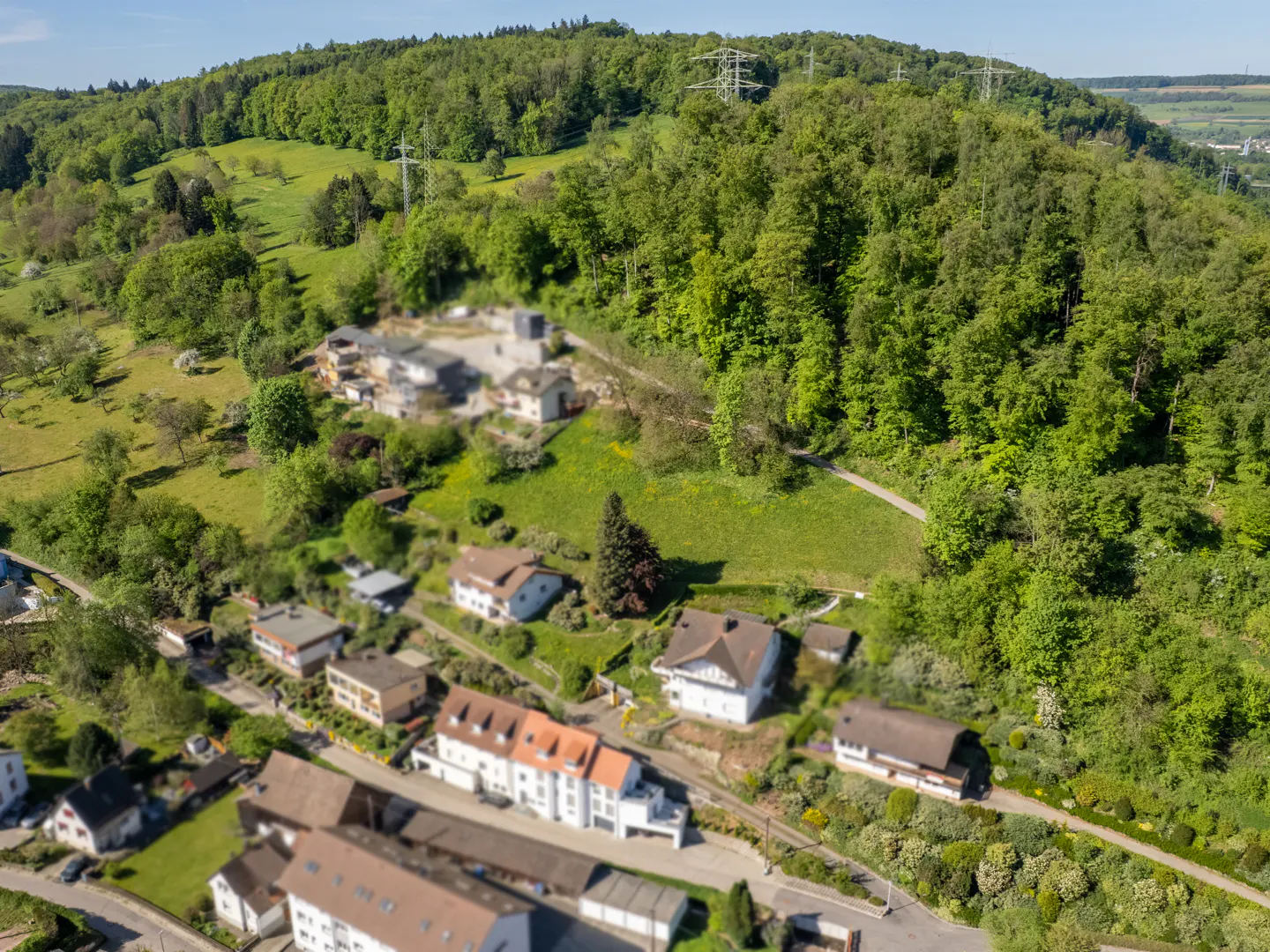 Aerial view of houses nestled in a green, hilly landscape with dense trees and a clear blue sky.