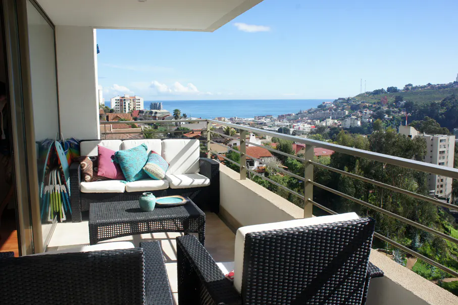 Balcony with black wicker furniture, white cushions, and colorful pillows. Ocean and city views beyond the railing.