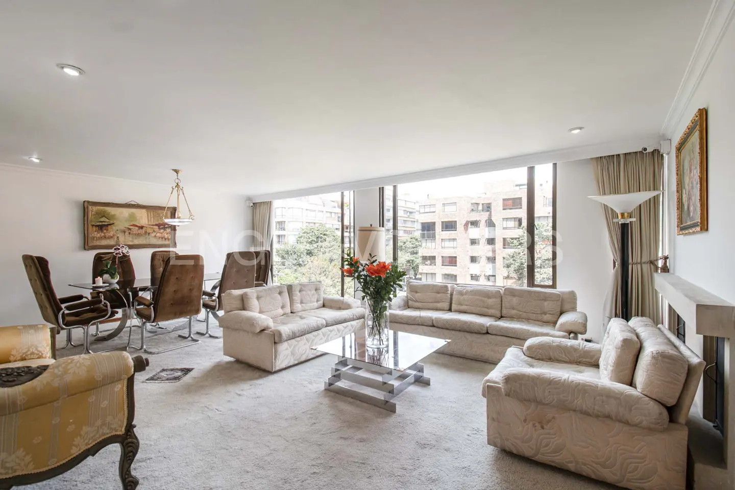 Bright living room with beige sofas, glass table, and large windows overlooking city buildings. Dining table with brown chairs in the background.
