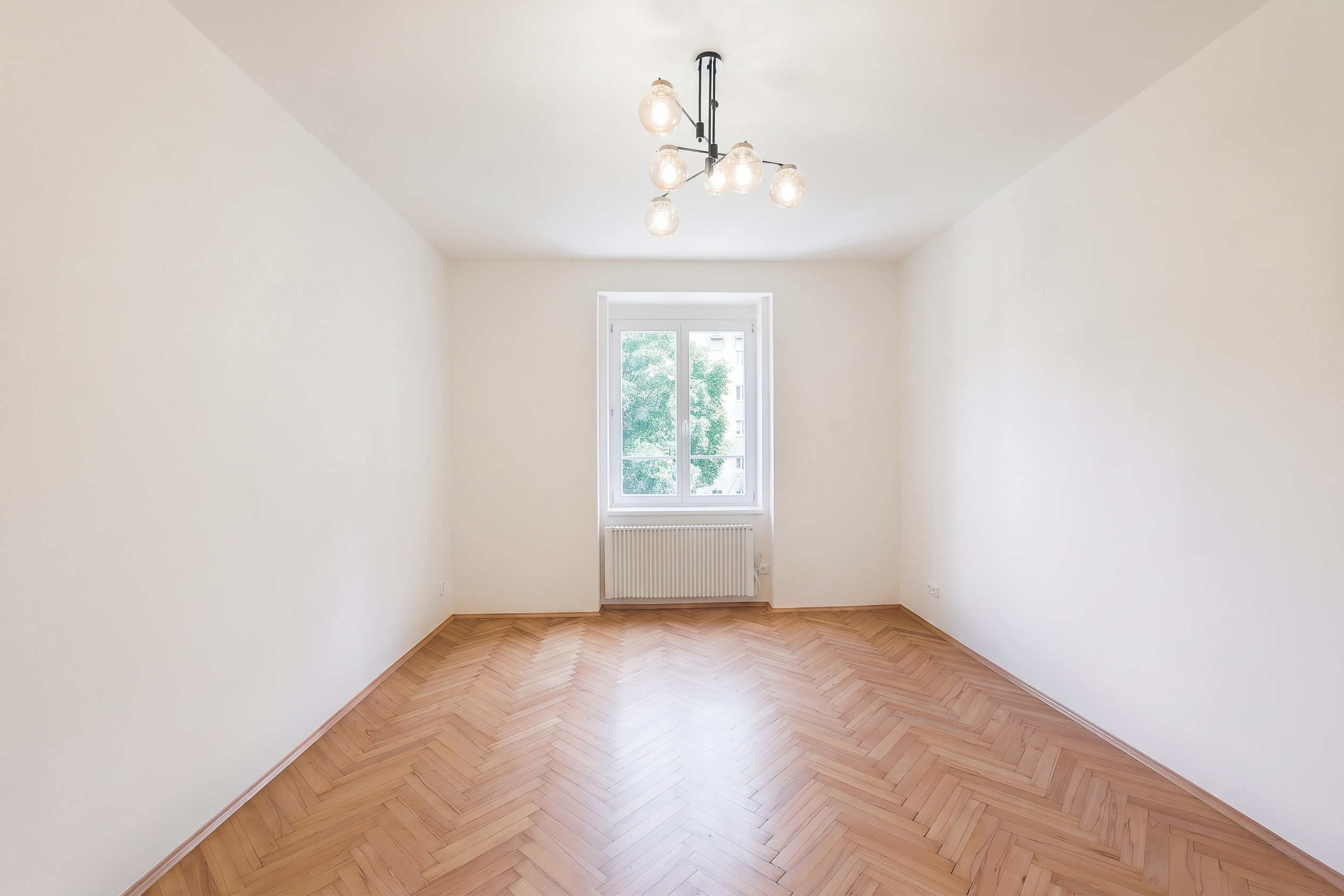 Empty room with white walls, light wood herringbone floors, a modern chandelier, and a window with a radiator below.