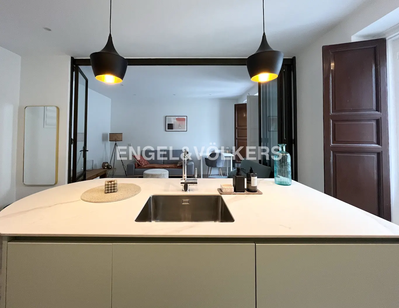 Open-concept kitchen with a white countertop island, stainless steel sink, and black pendant lights. Living room with gray sofa in the background.