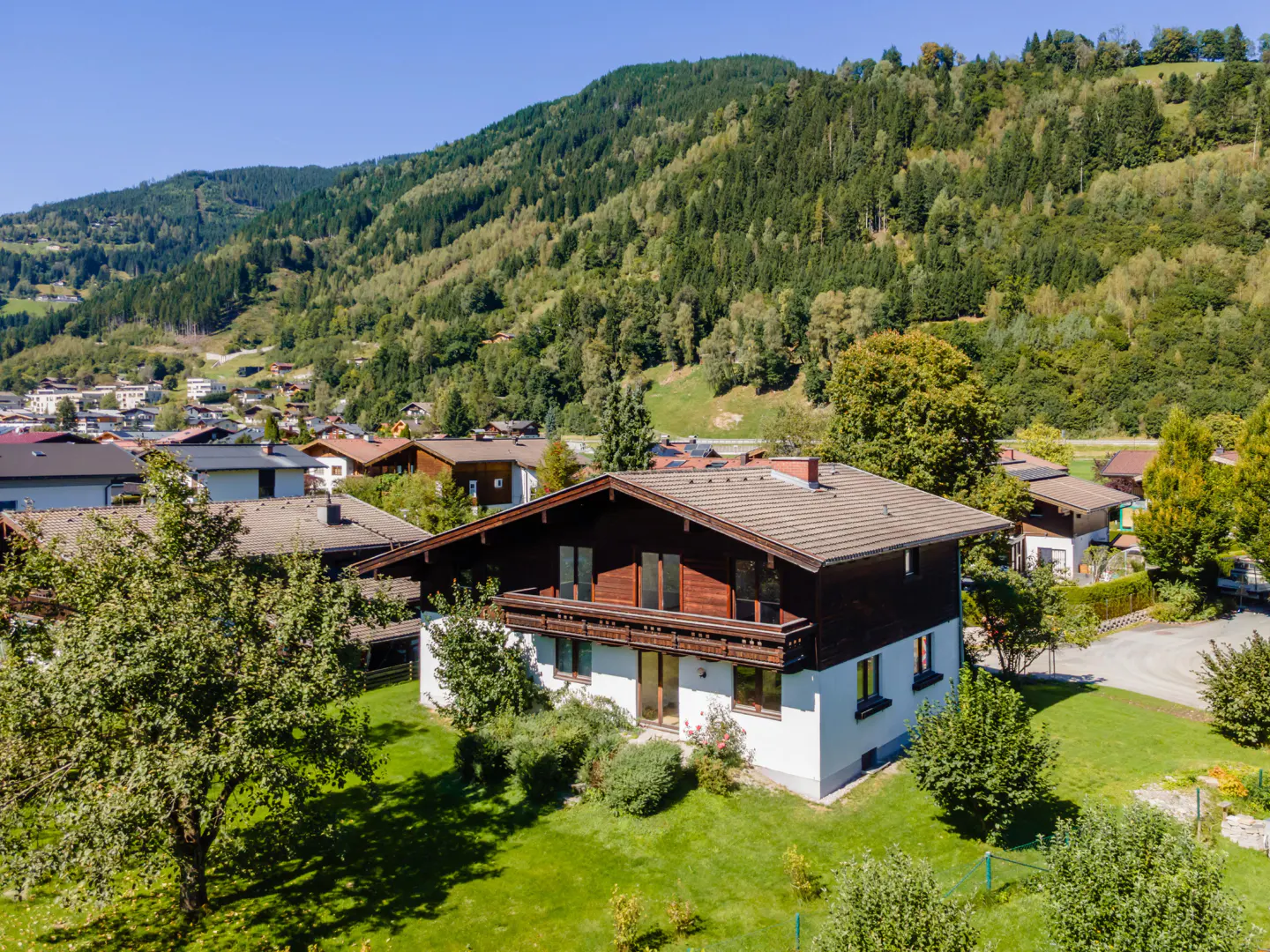 Aerial view of a two-story white house with a dark brown roof, surrounded by green lawn and trees, set against a mountain backdrop.