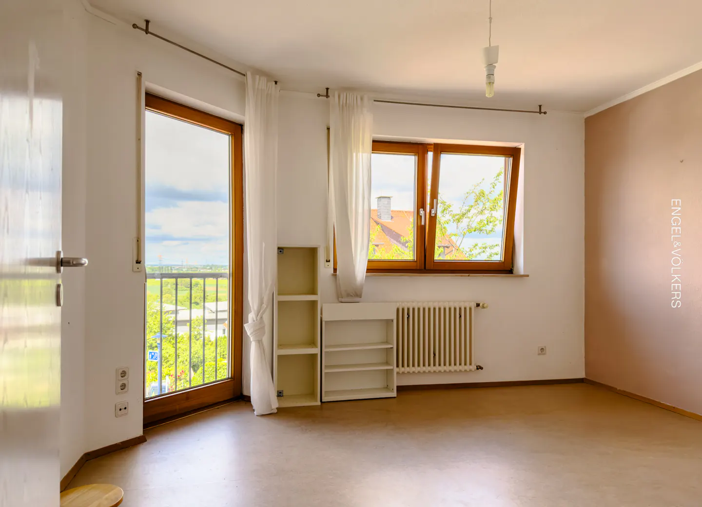 Bright, empty room with wood-framed windows and door to balcony. White walls, beige floor, radiator, and white shelving units. Natural light.