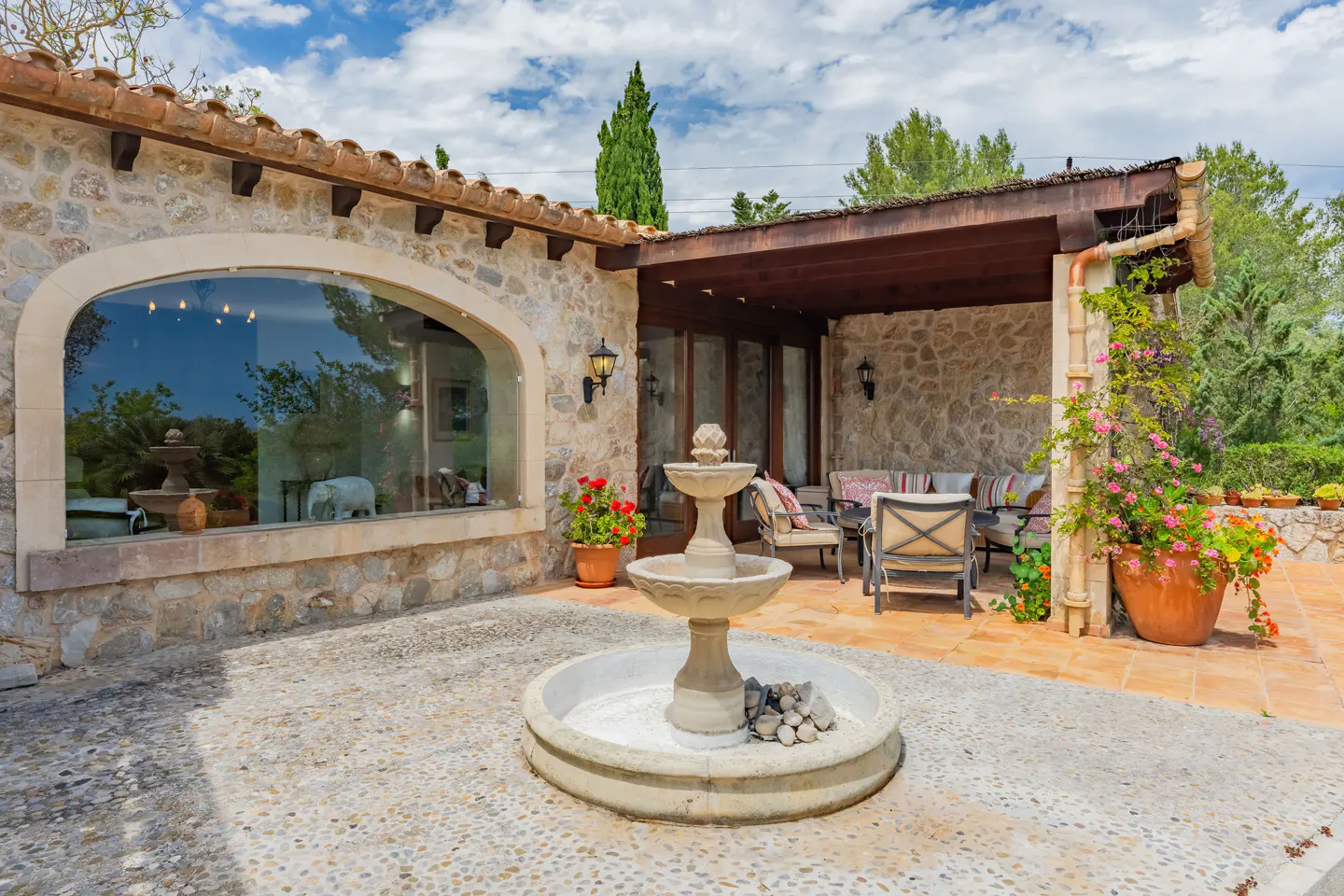 Stone patio with a tiered fountain in the foreground. Seating area under a covered porch, and a large arched window on the left.