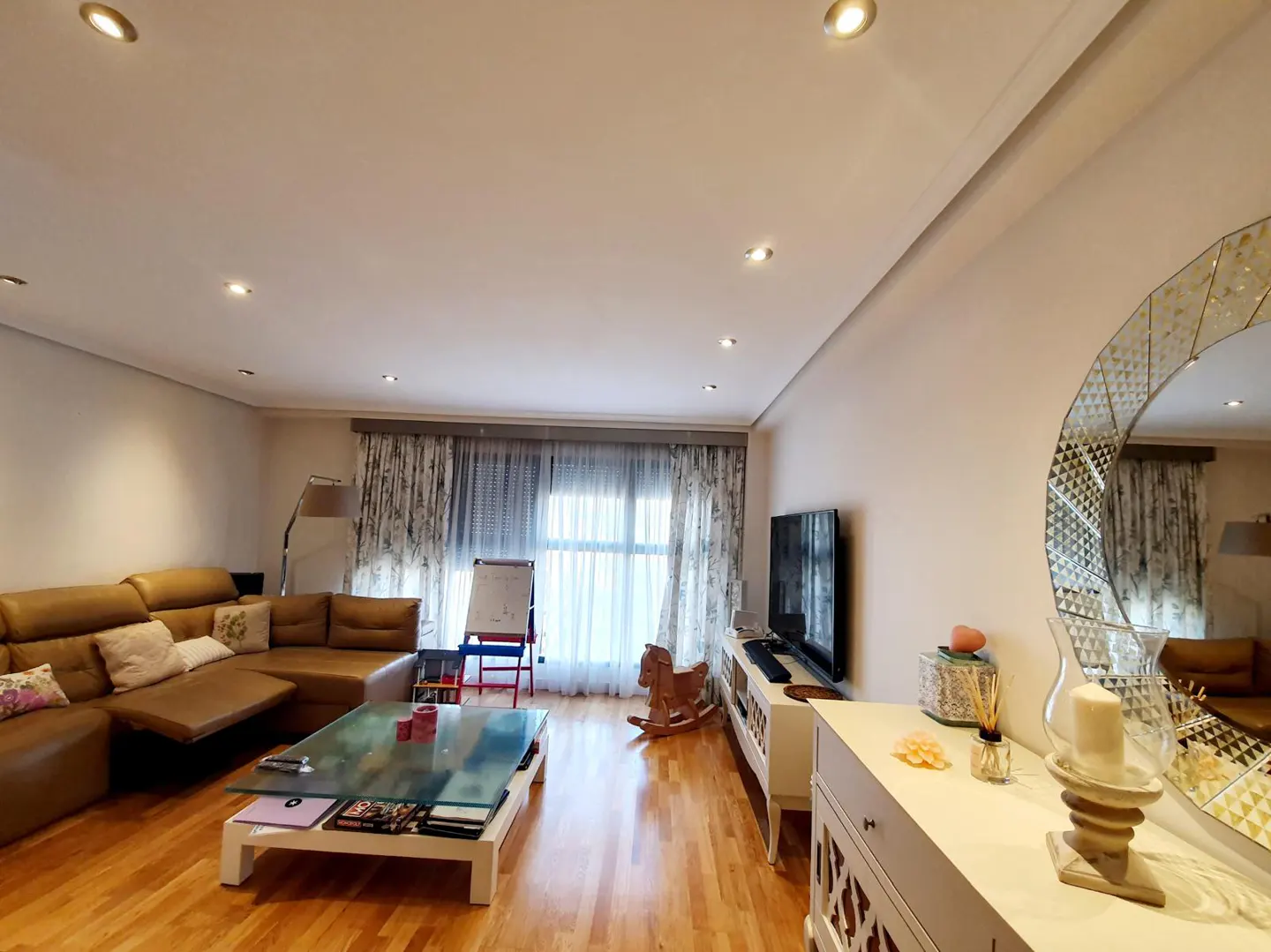Bright living room with hardwood floors, a brown leather sectional sofa, a glass coffee table, and a large round mirror.