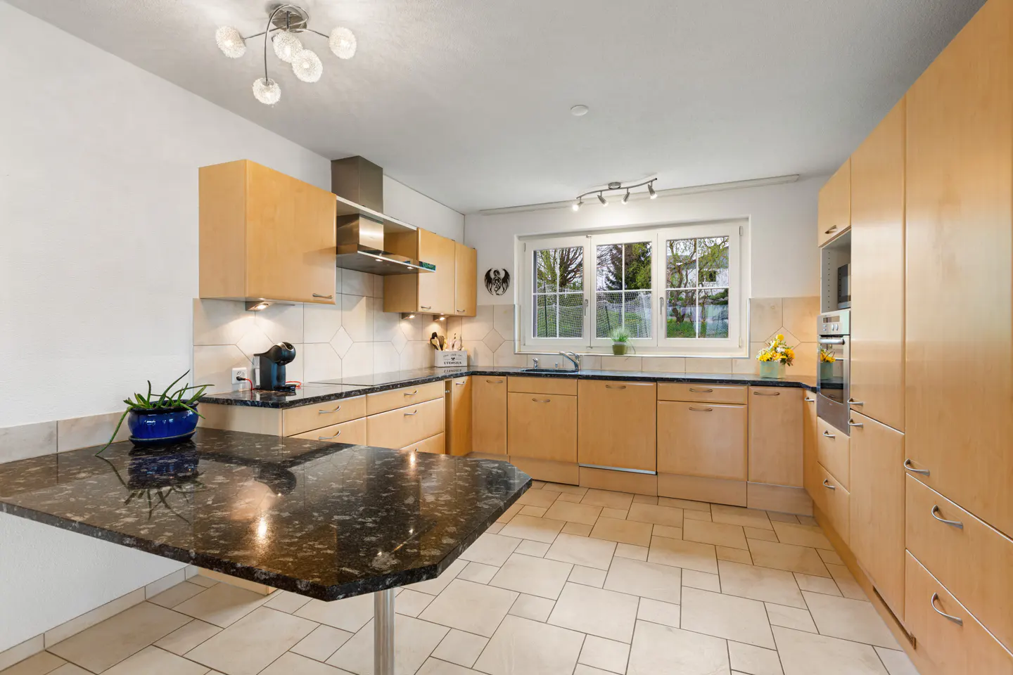 Bright kitchen with light wood cabinets, black granite countertop, and tile flooring. A window overlooks a green yard.