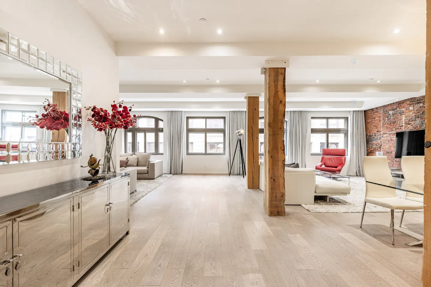 Open-concept living space with hardwood floors, exposed brick, and large windows. A mirrored console table and red floral arrangement.