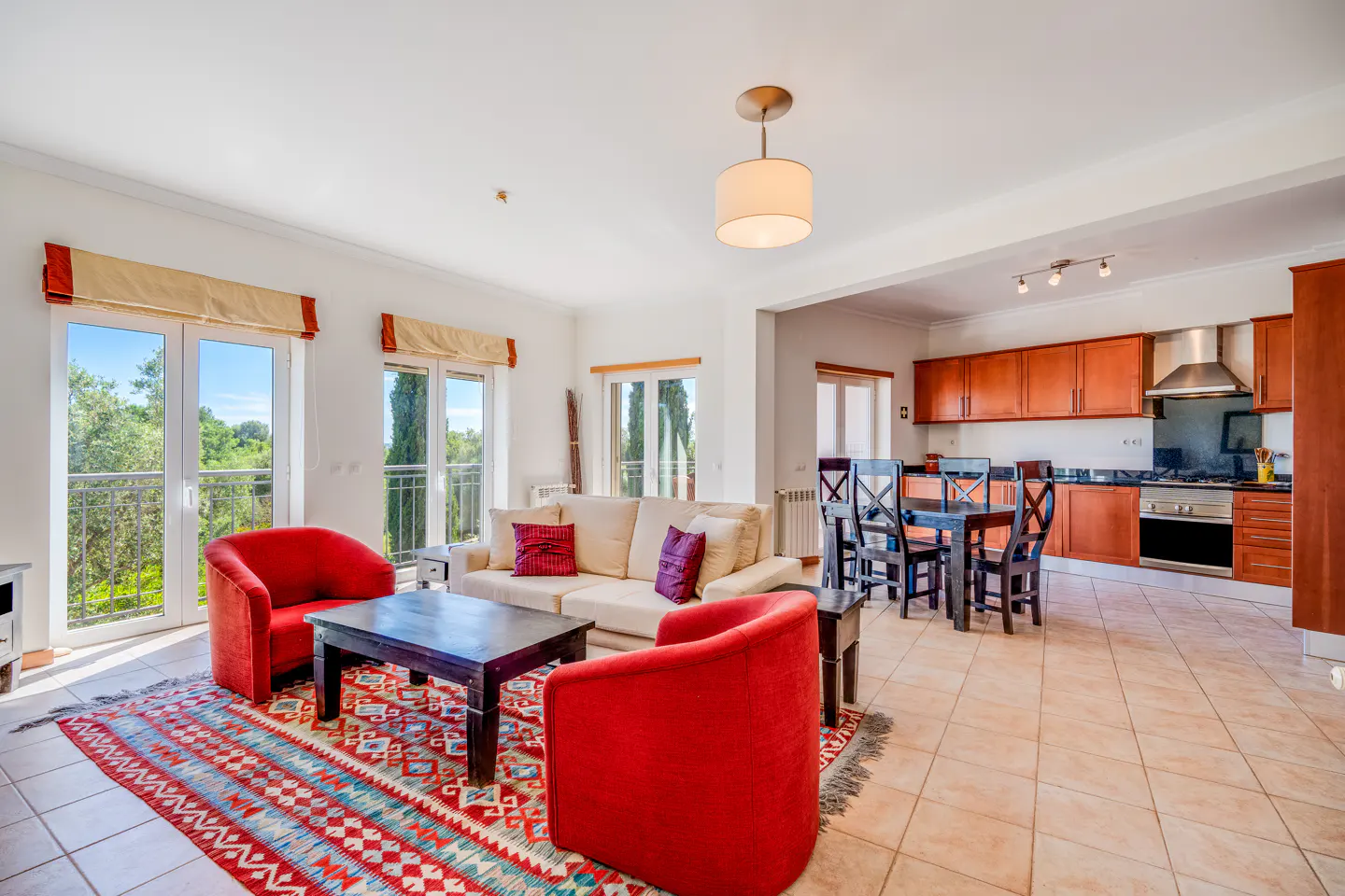 Bright living room with red chairs, a beige sofa, and a colorful rug. The kitchen and dining area are visible in the background.