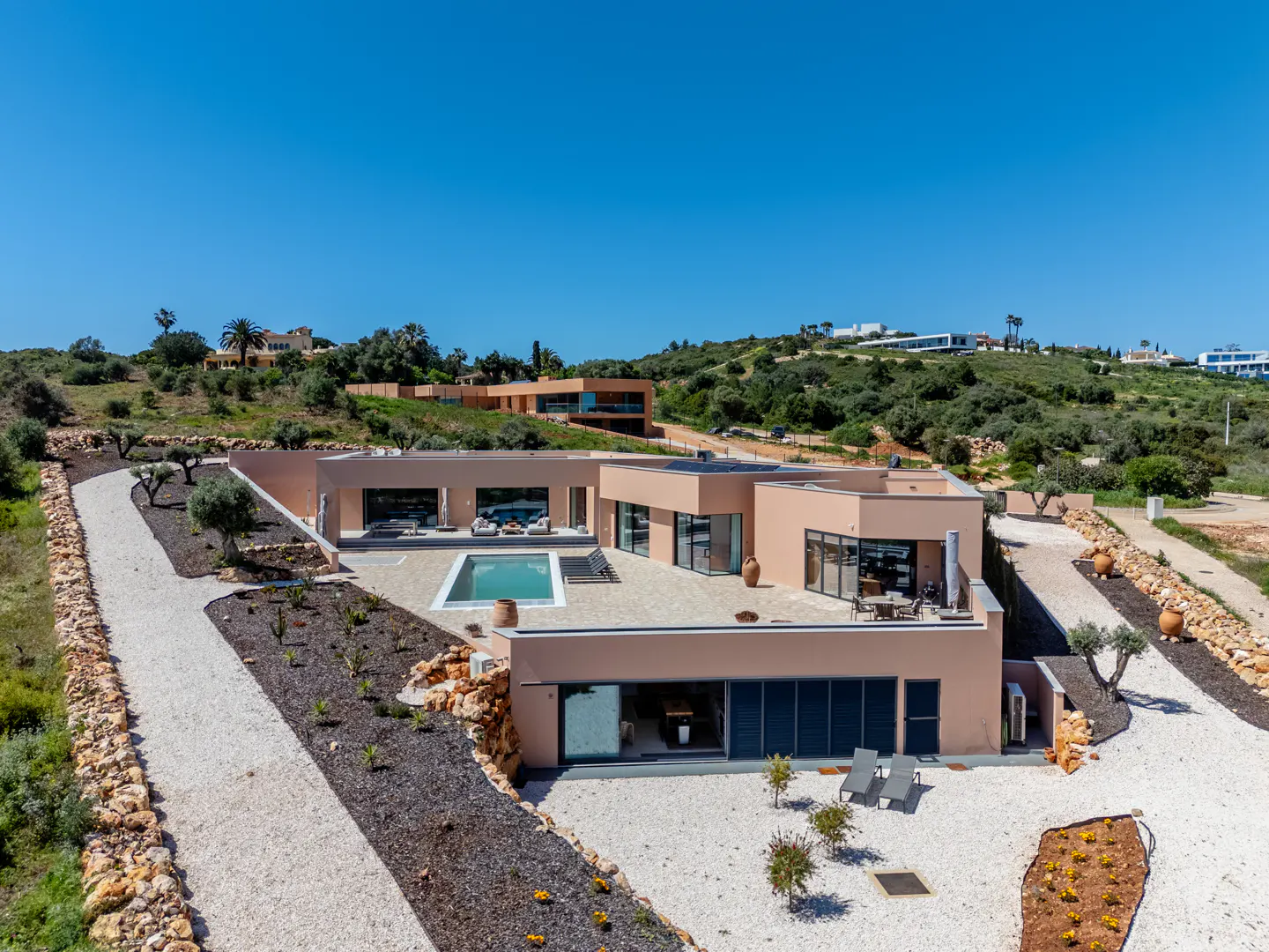 Aerial view of a modern, single-story peach-colored house with a pool, patio furniture, and gravel landscaping under a clear blue sky.