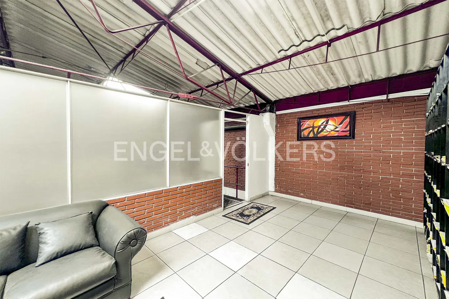 Interior view of a room with a gray sofa, brick walls, white tile floor, and a metal roof structure. An abstract painting hangs on the brick wall.