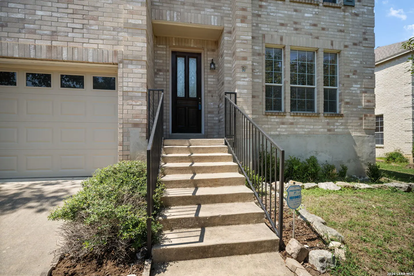 Exterior view of a two-story brick house with a beige garage door and a black front door with glass panels. Concrete steps lead to the entrance with black metal railings. Windows with blinds are visible.