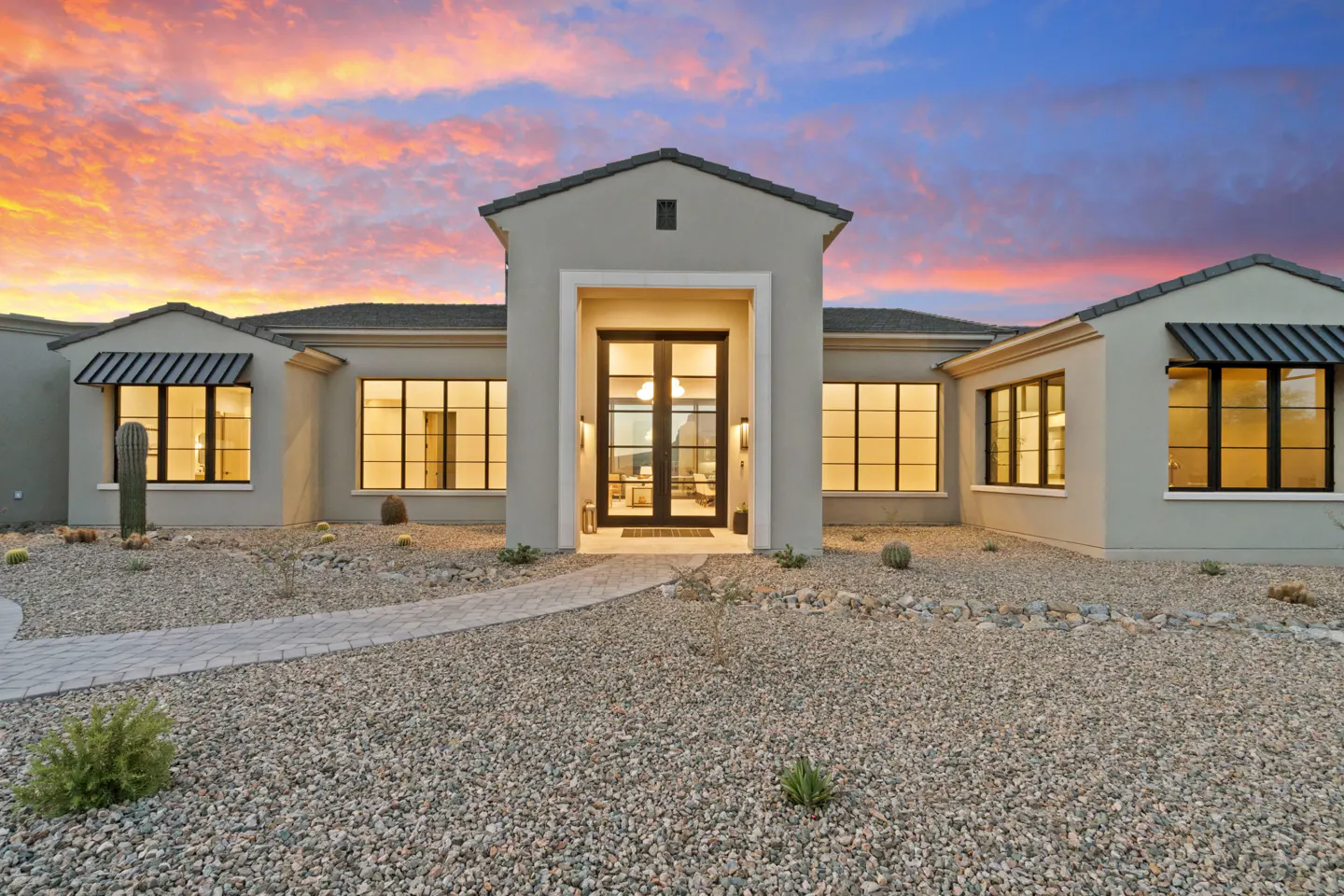 Modern desert home with a stone walkway, gravel landscaping, and large windows under a vibrant sunset sky.