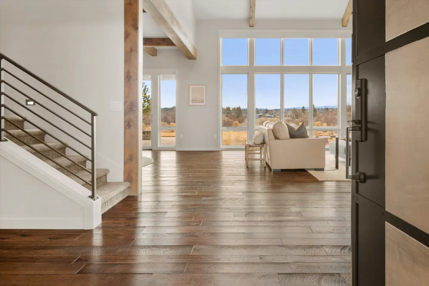Open front door view of a modern home interior with hardwood floors, stairs, and a large window overlooking a scenic landscape.