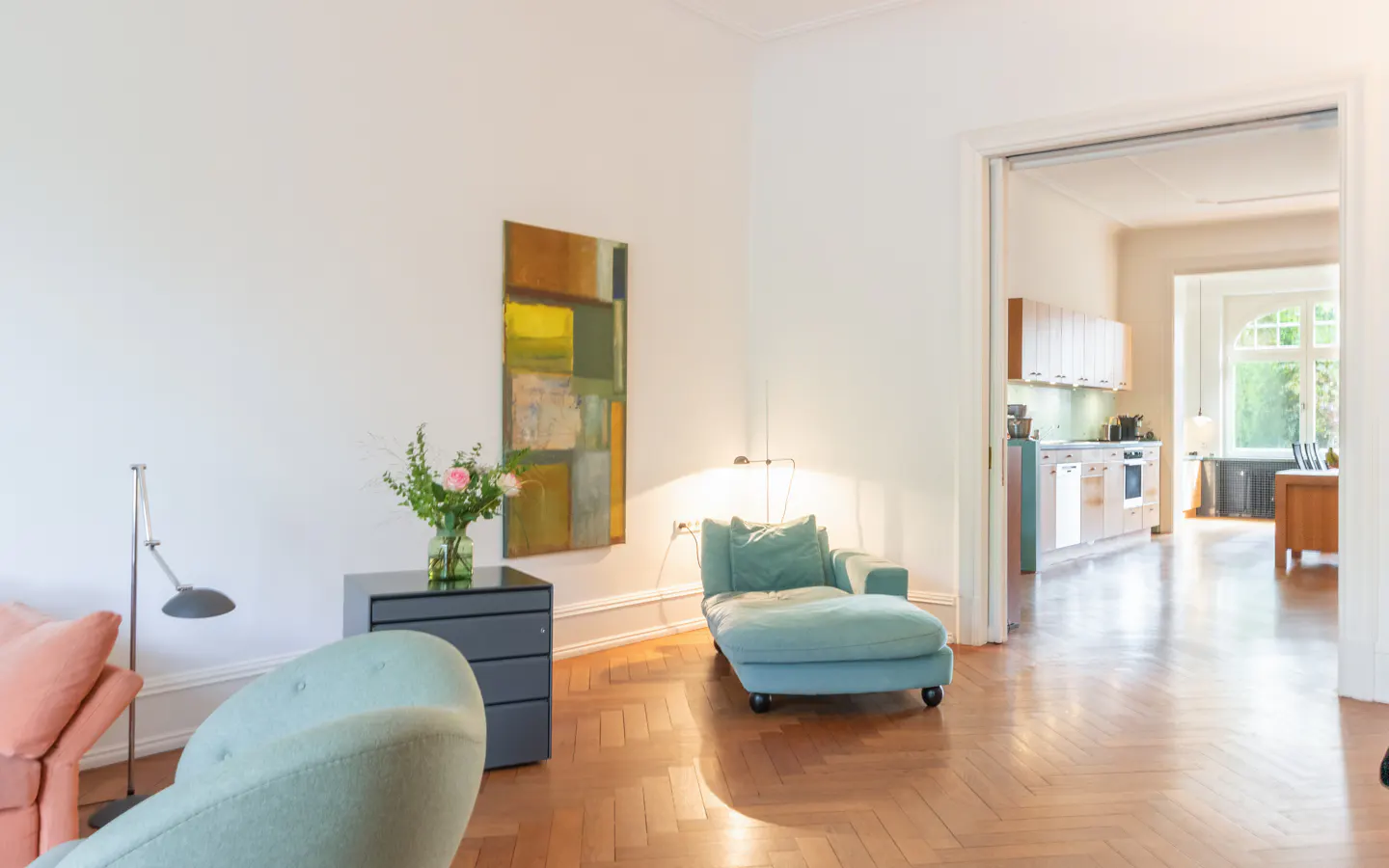 Bright living room with parquet floors, a blue chaise lounge, and a view into the kitchen and dining area.