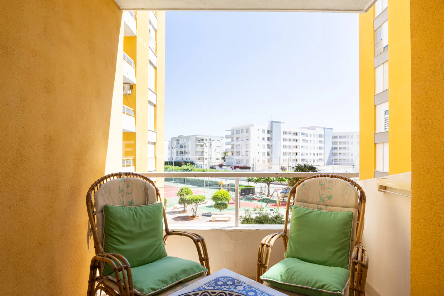 Balcony view with two wicker chairs, green cushions, and a blue-tiled table. Outside, there are buildings, a tennis court, and a playground.