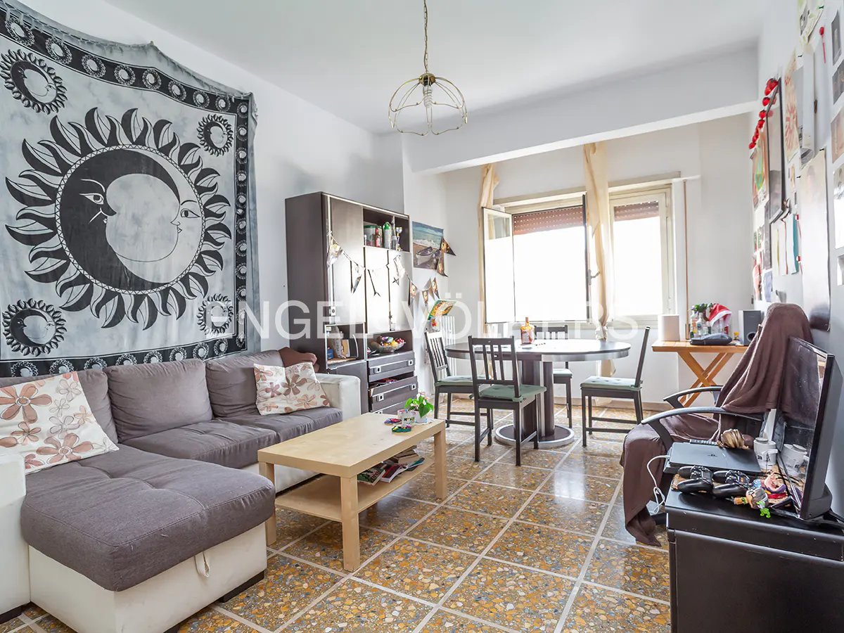 A living room with a gray sectional sofa, a wooden coffee table, and a dining table with chairs. A tapestry with a sun and moon design hangs on the wall.
