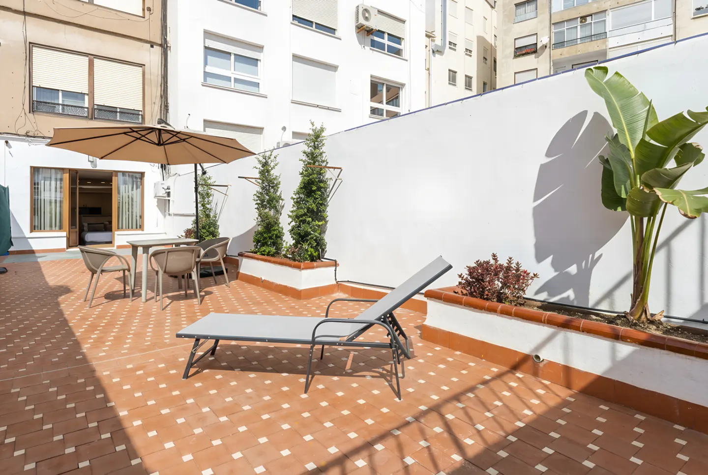 Outdoor patio with terracotta tile, white walls, and potted plants. A lounge chair and table with chairs sit under a tan umbrella.