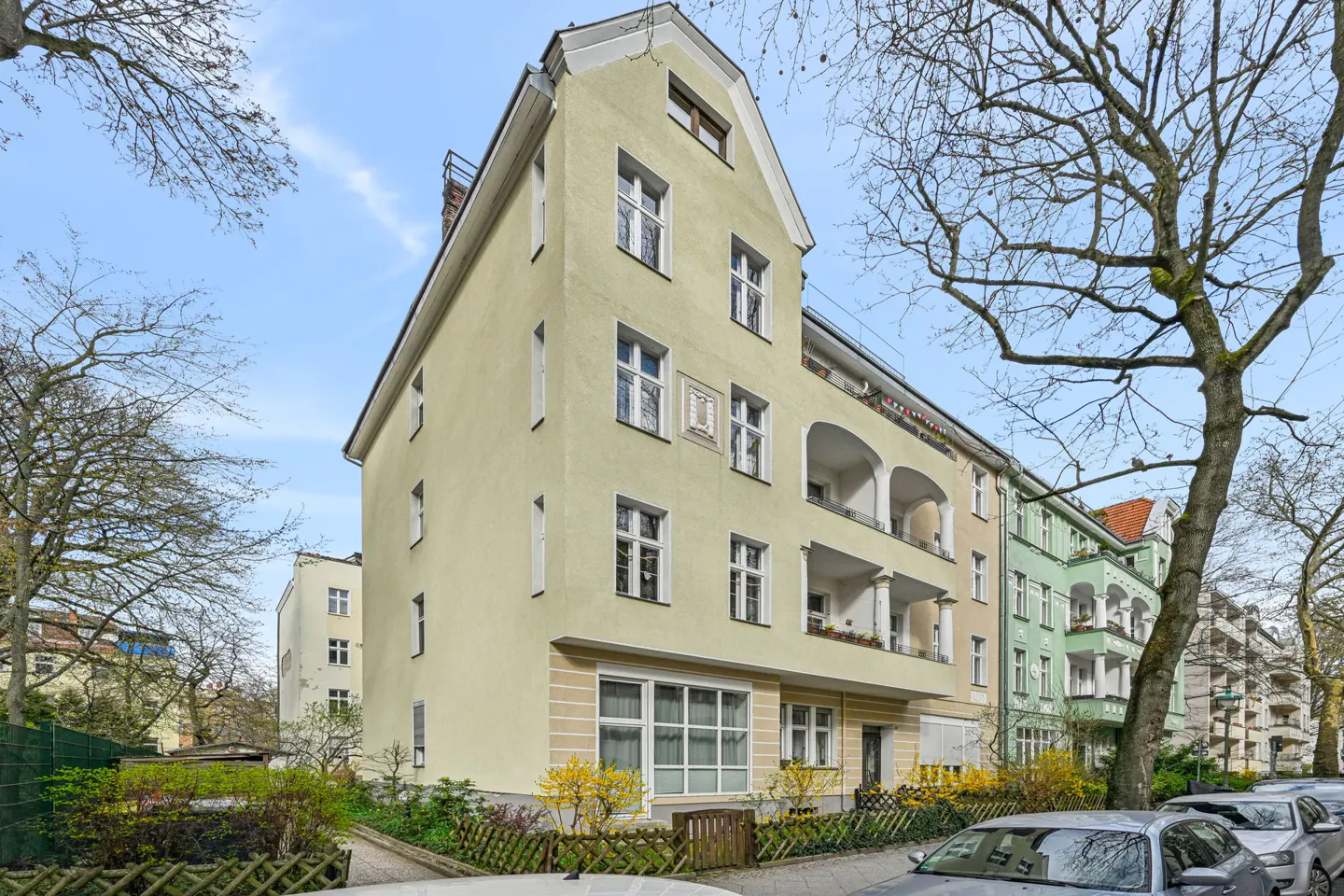 A three-story, light yellow apartment building with white trim and balconies, framed by trees and parked cars.