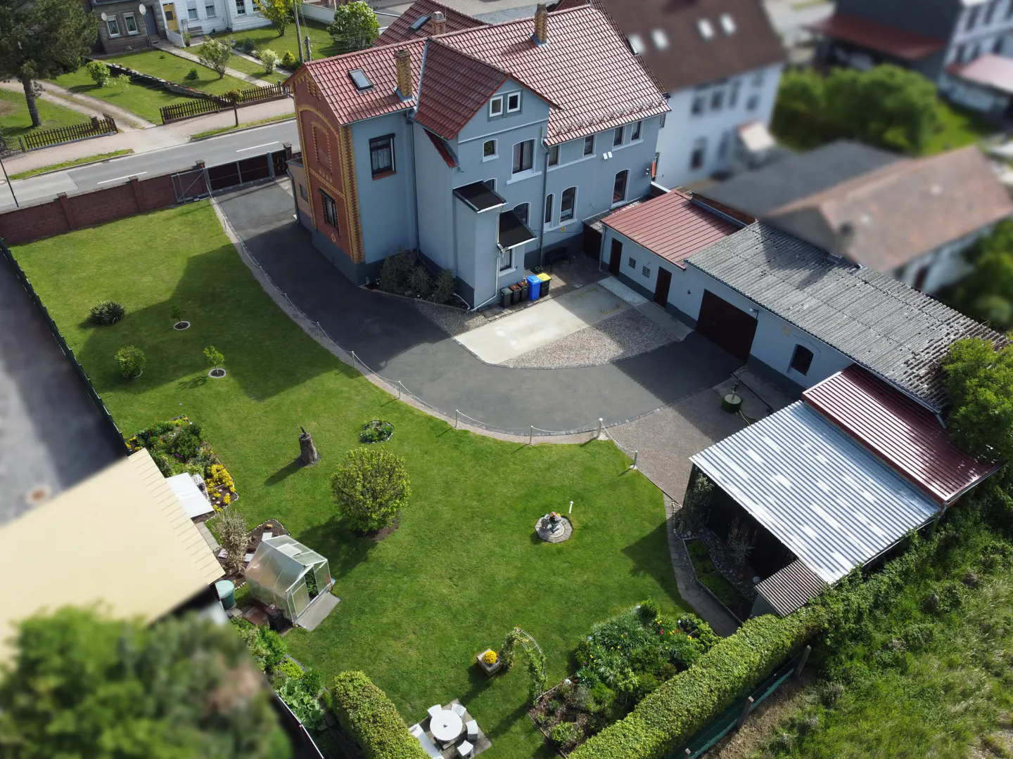 Aerial view of a grey house with a red roof, large green lawn, paved driveway, and a small greenhouse in the backyard.