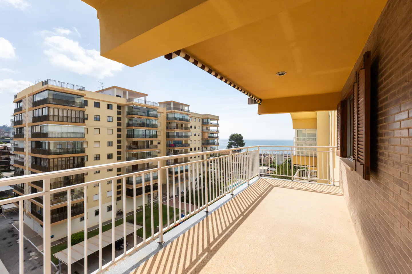 View from a balcony with white railings, overlooking apartment buildings and the ocean on a sunny day.