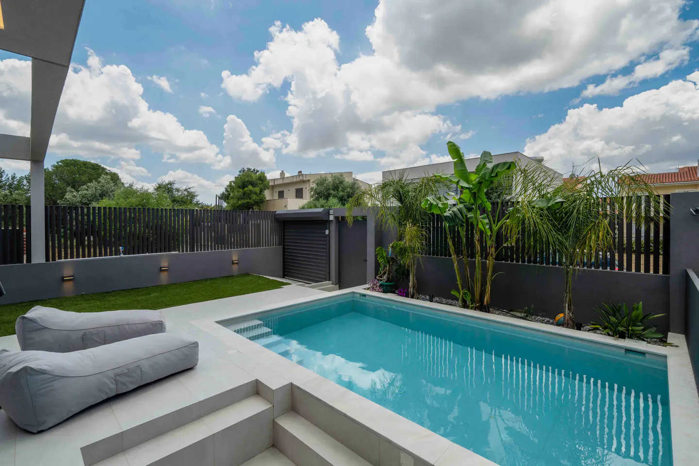 A backyard with a pool, two gray beanbag chairs, a gray fence, and green plants under a cloudy blue sky.