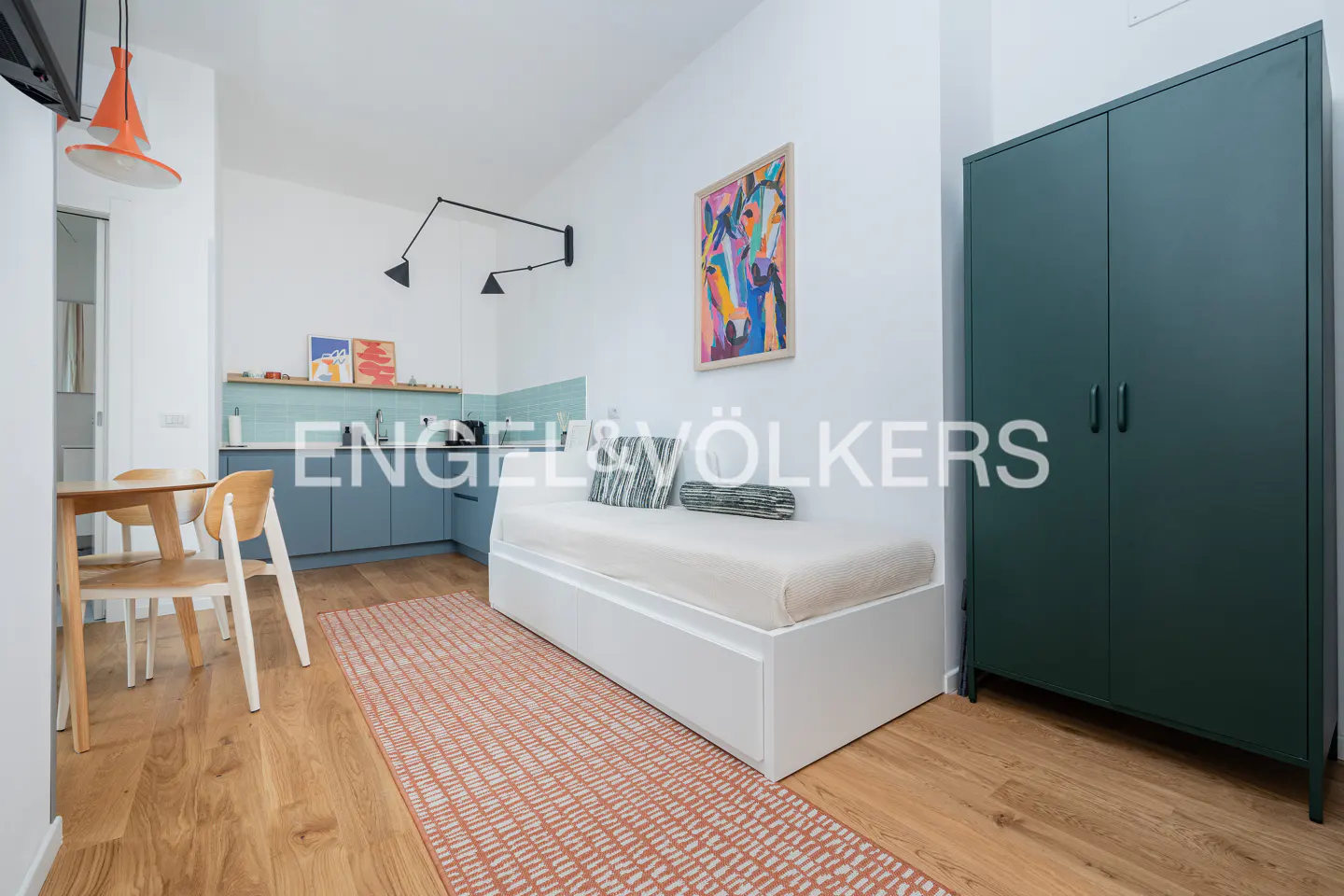 Modern studio apartment with a white daybed, dark green wardrobe, small dining area, and a patterned orange rug.