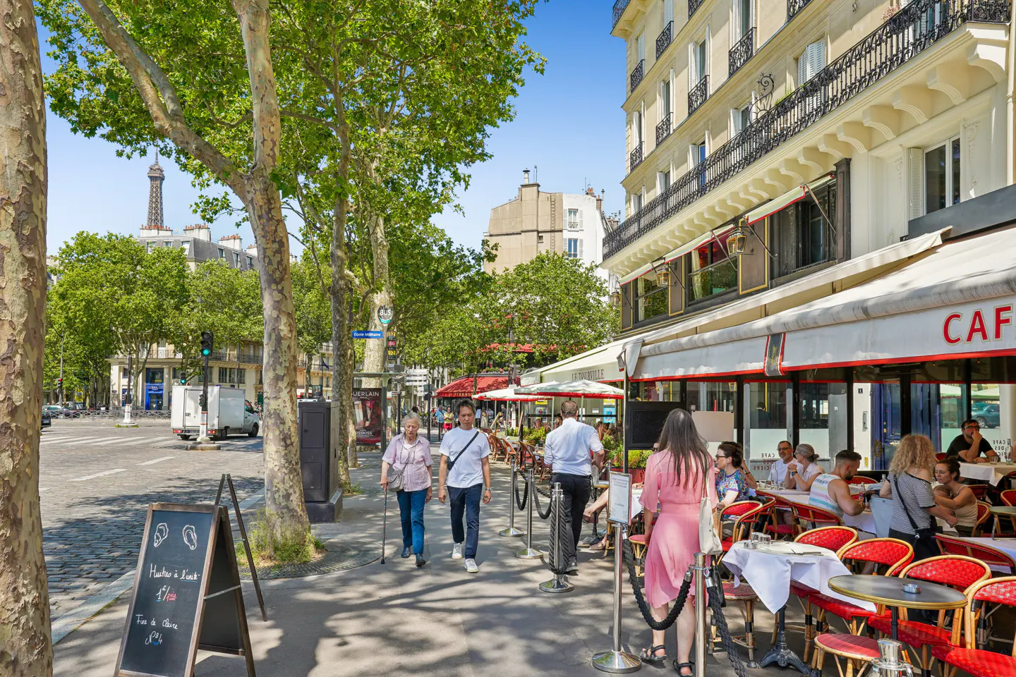 Paris street scene with sidewalk cafe, people dining, and the Eiffel Tower visible in the distance.