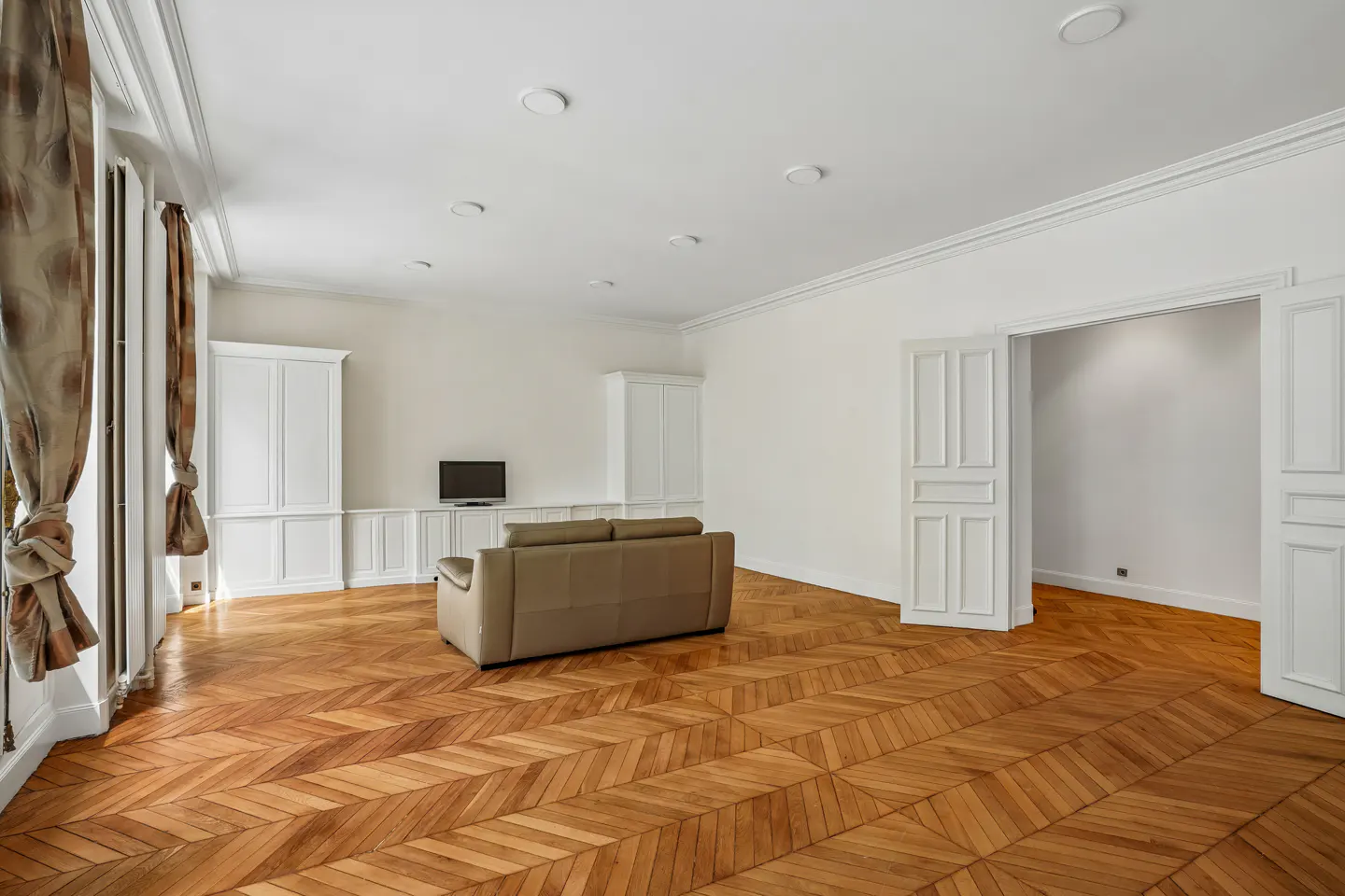Bright, empty living room with parquet floors, white walls, and a tan sofa. Curtains frame a window to the left. A TV sits on a built-in cabinet.