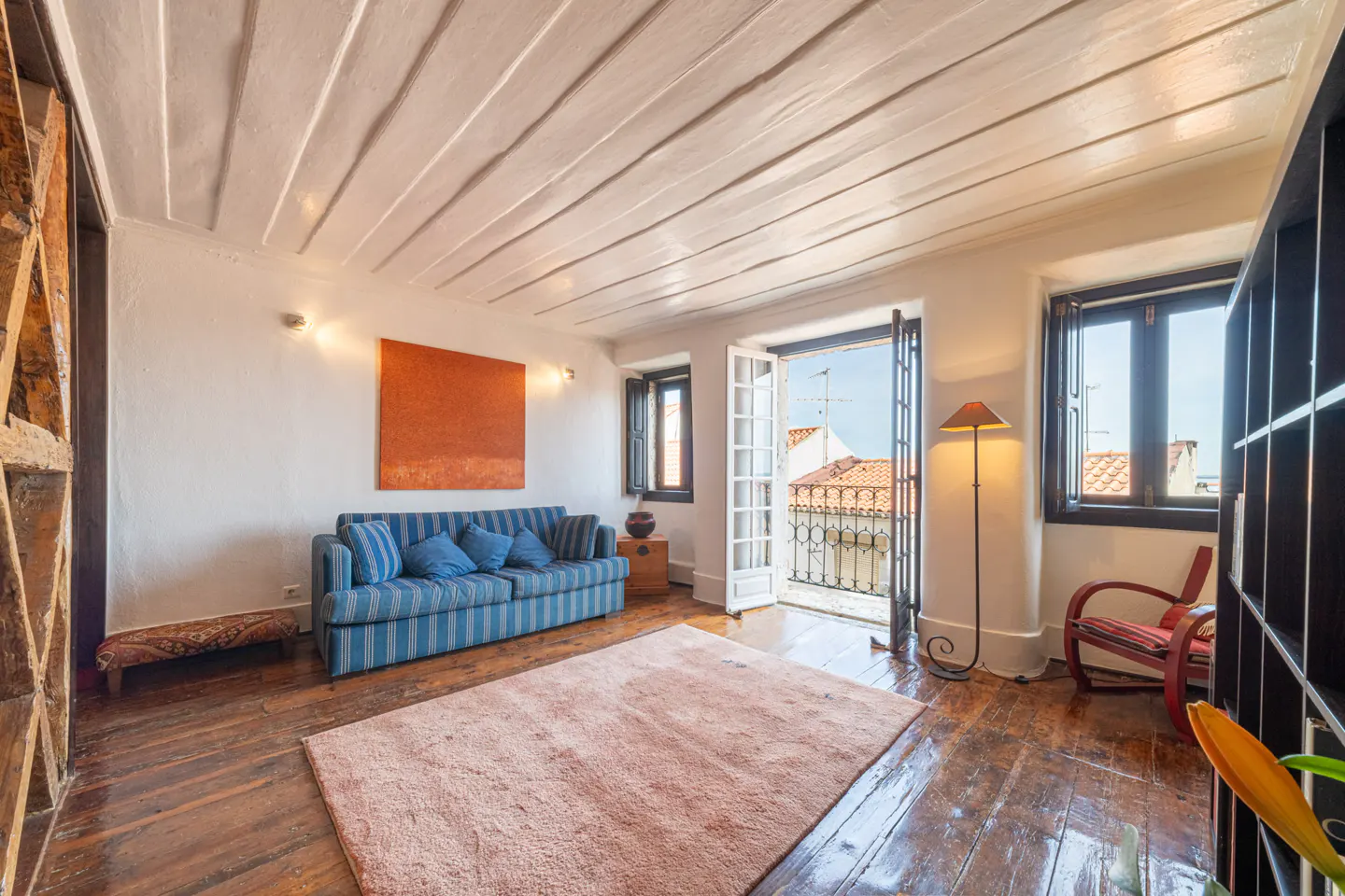 Bright living room with wood floors, white ceiling, and balcony. A blue striped sofa sits under an orange painting.