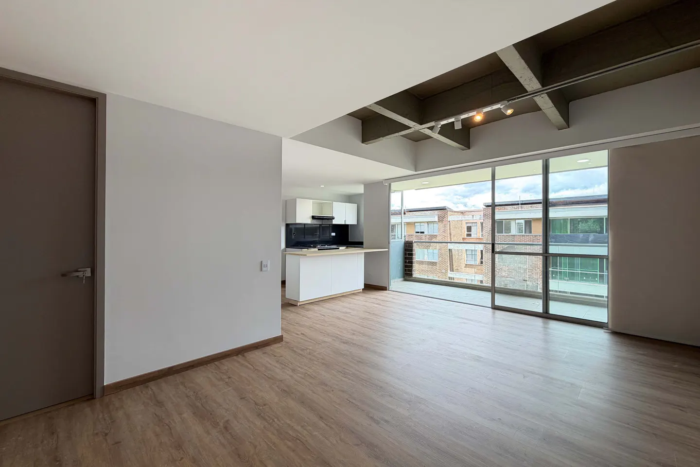 An open-concept apartment with wood floors, gray walls, and a white kitchen. A large window overlooks a brick building.