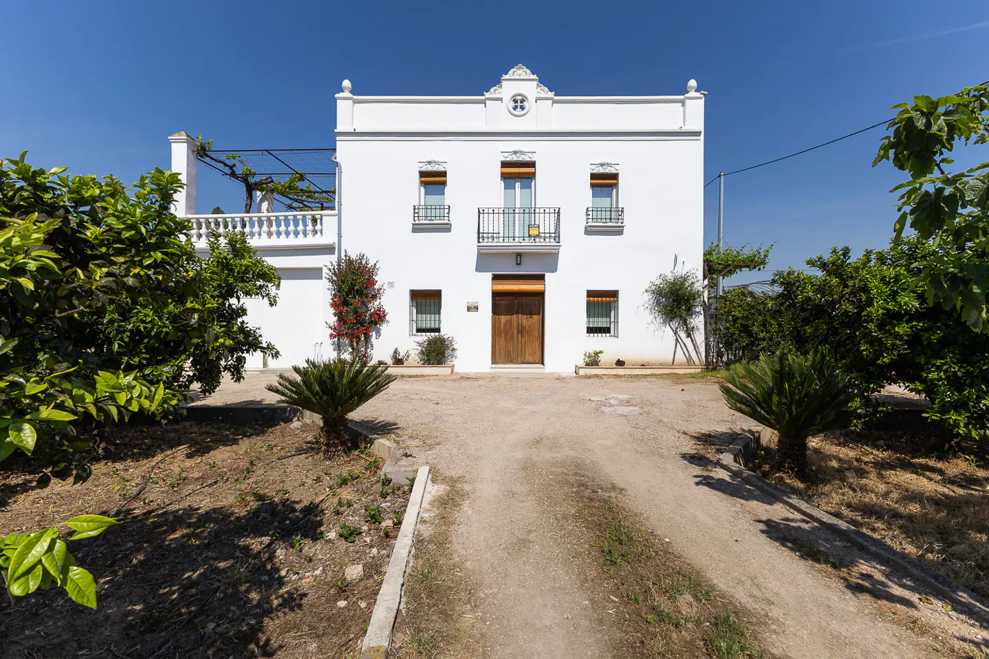 White two-story house with a brown wooden door and small balconies under a clear blue sky. Green trees surround the house.