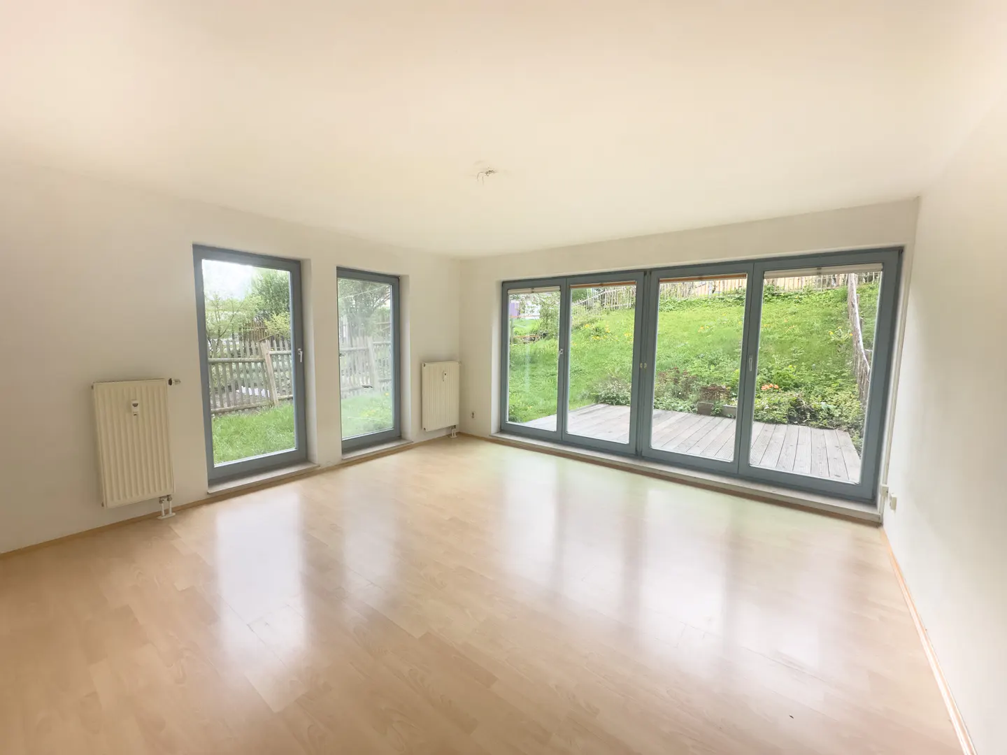 Empty room with light wood floors, white walls, and large glass doors opening to a green backyard and wooden deck.