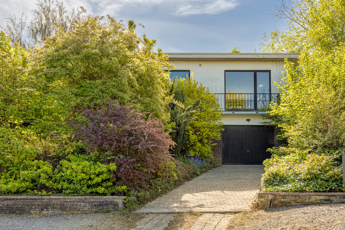Exterior of a white, two-story house with a black garage door and balcony, surrounded by lush green and purple bushes.