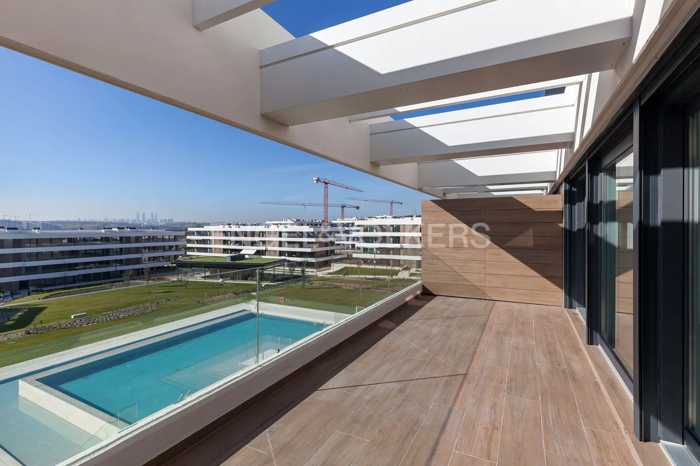 Balcony view of a modern apartment complex with a pool, green lawn, and city skyline under a blue sky. White pergola above.