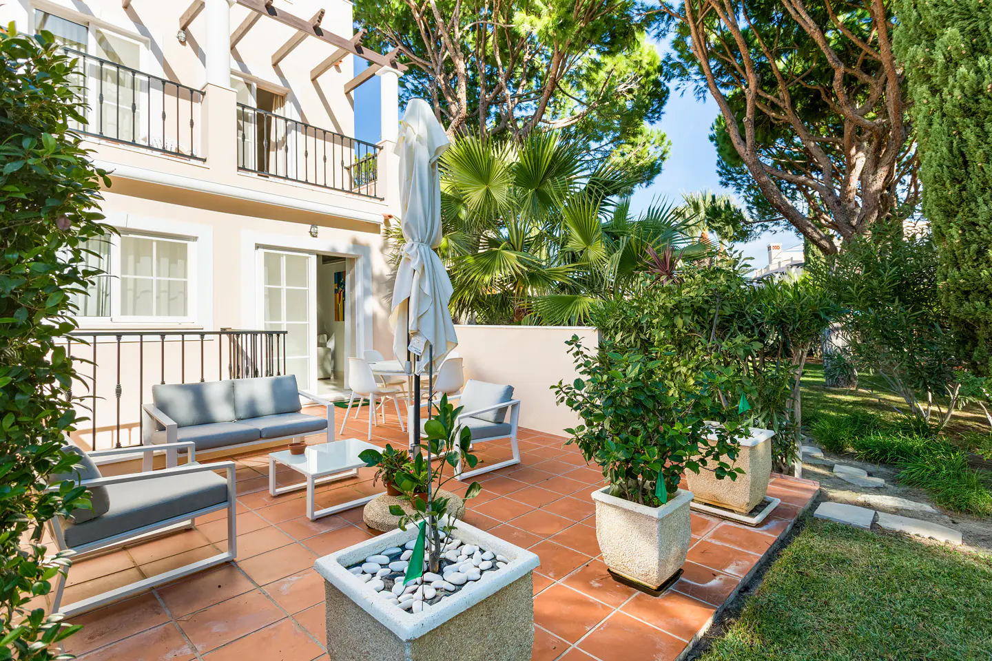 Outdoor patio with gray furniture, terracotta tiles, and lush greenery. A white umbrella stands near a table and chairs.