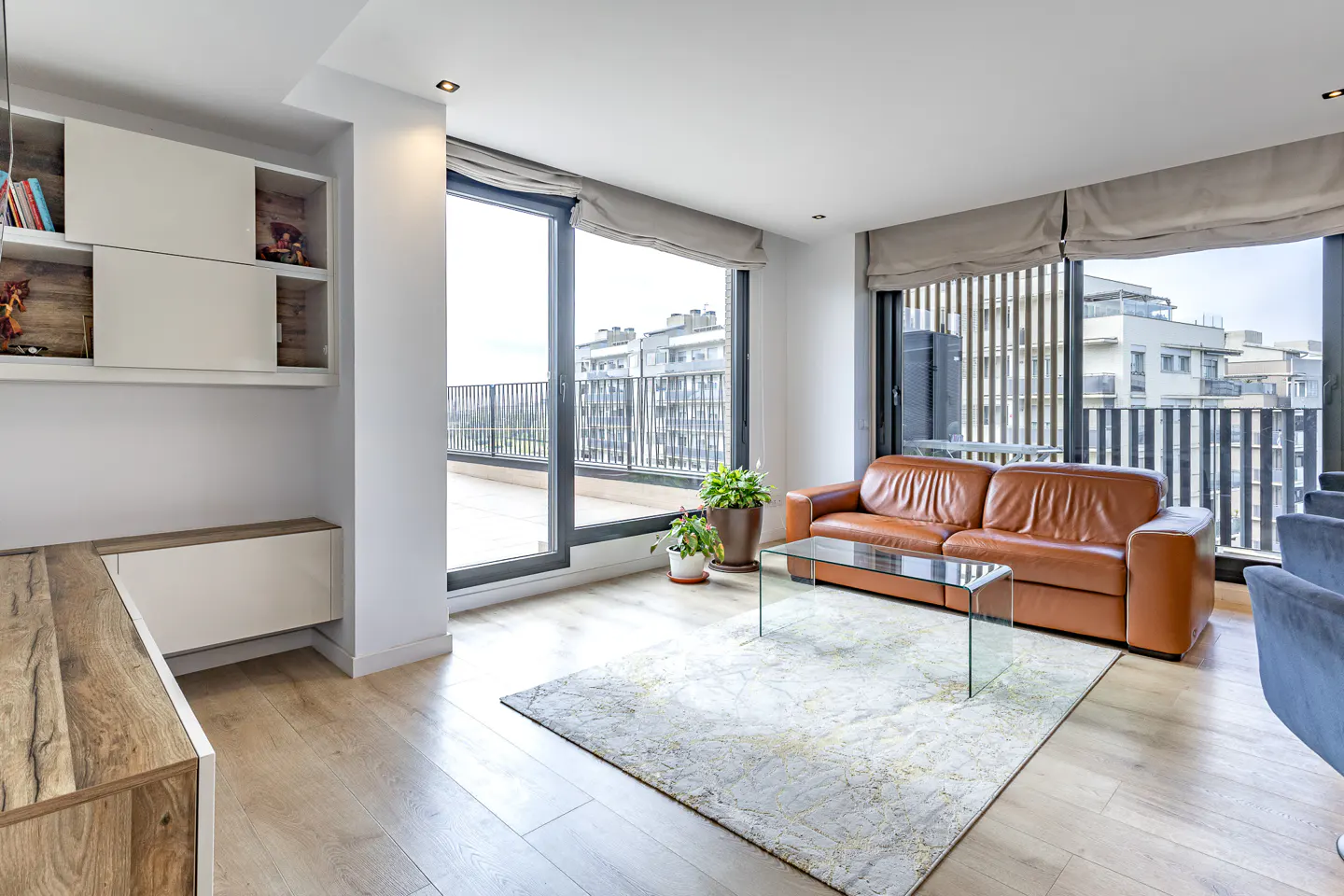 Bright living room with a brown leather sofa, glass table, and light wood floors. Large windows offer a city view.