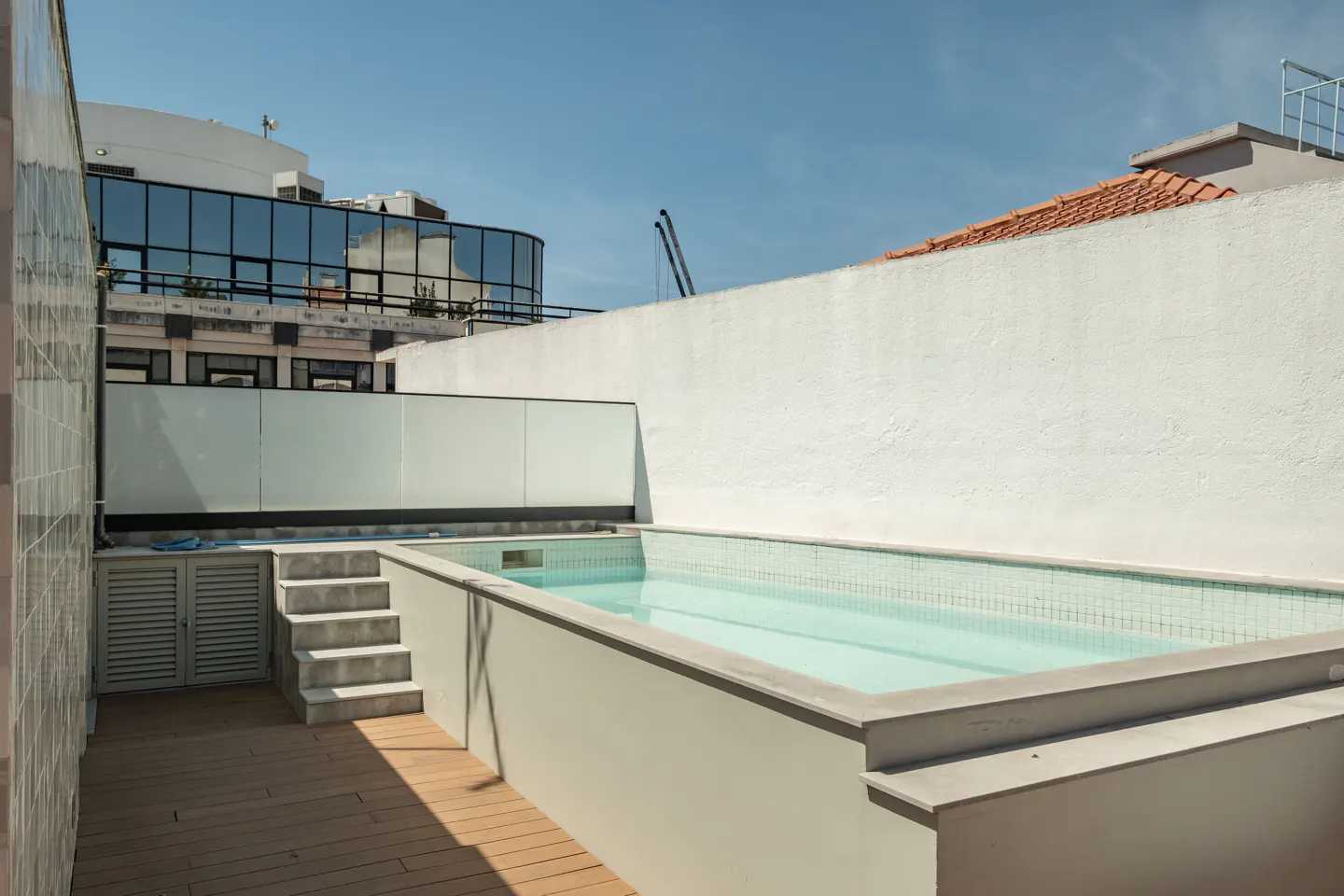 Rooftop swimming pool with light blue water, wooden decking, concrete steps, and a glass railing against a white wall.
