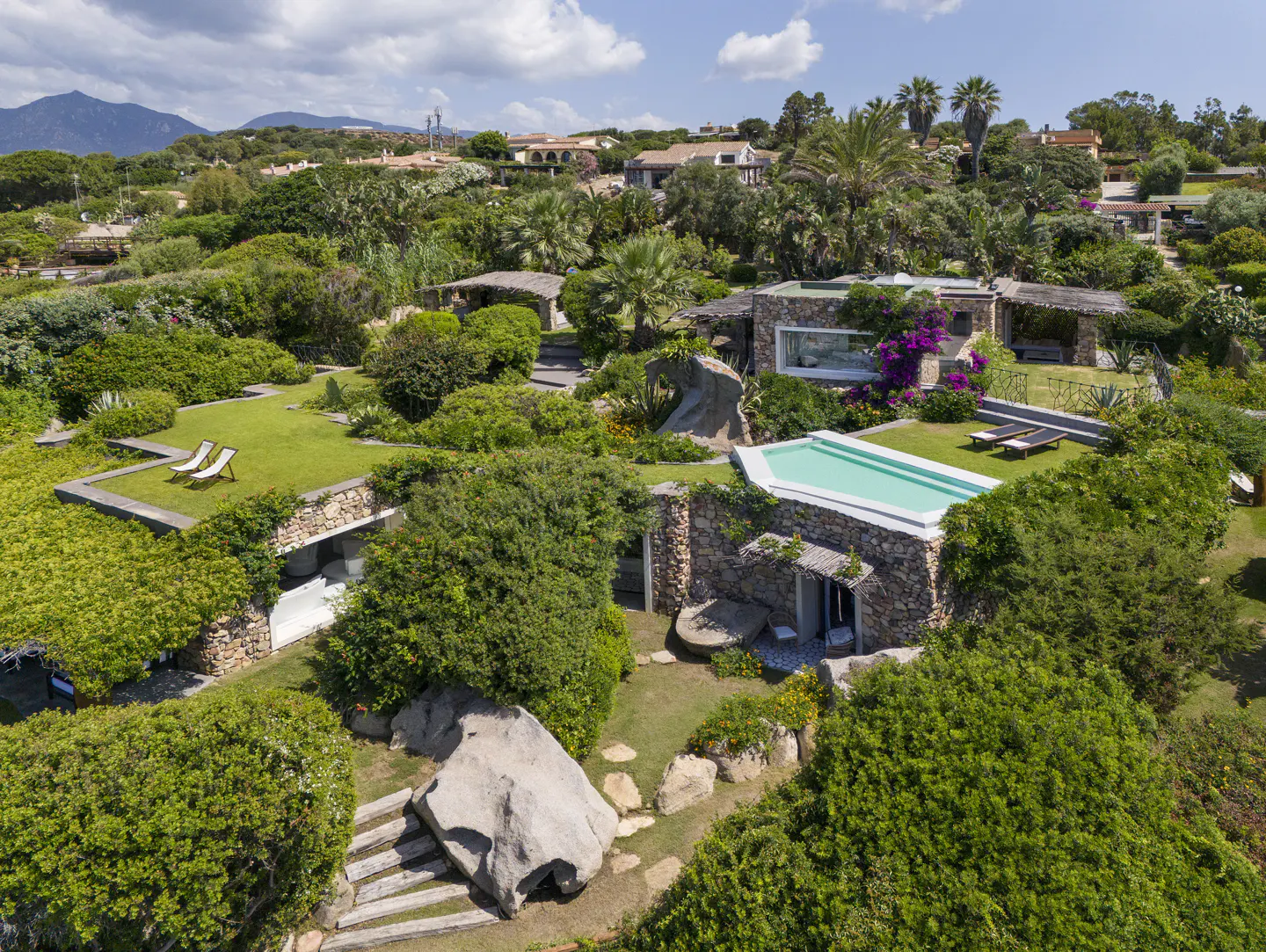 Aerial view of a stone house with a pool, surrounded by lush green vegetation and trees under a blue sky.