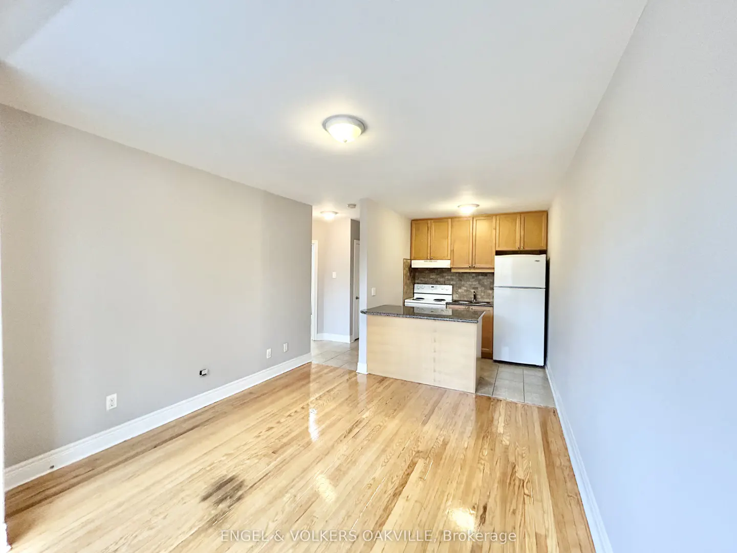 A bright apartment with hardwood floors, light gray walls, and an open kitchen with wood cabinets and a white refrigerator.