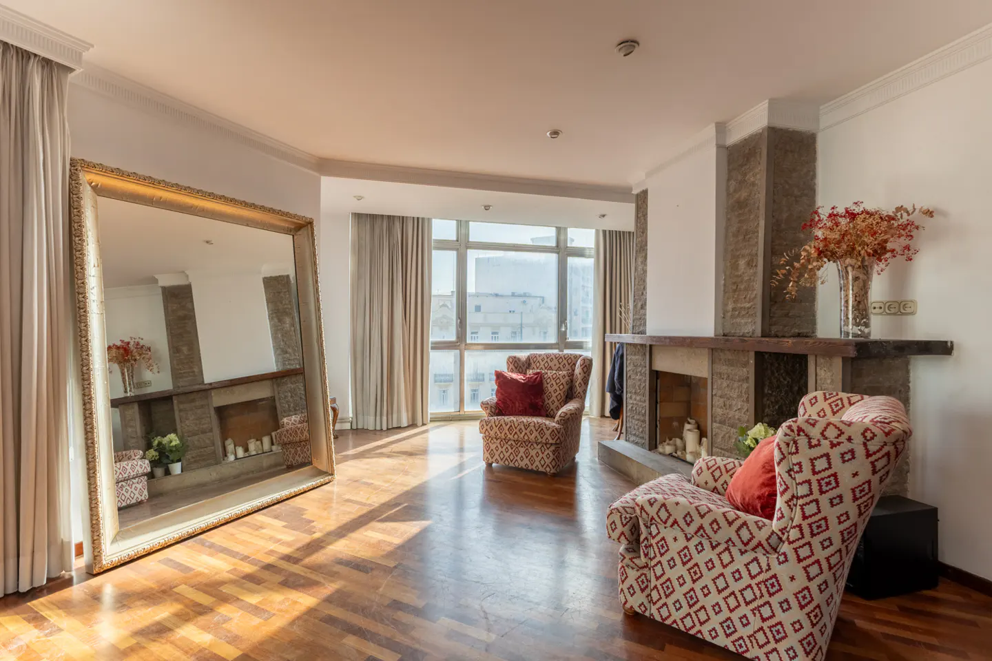 Bright living room with hardwood floors, large gold-framed mirror, patterned armchairs, and a stone fireplace. Curtains frame a window with a city view.