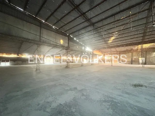 An empty warehouse with a gray concrete floor and a high, corrugated metal roof. Sunlight streams in from the sides.