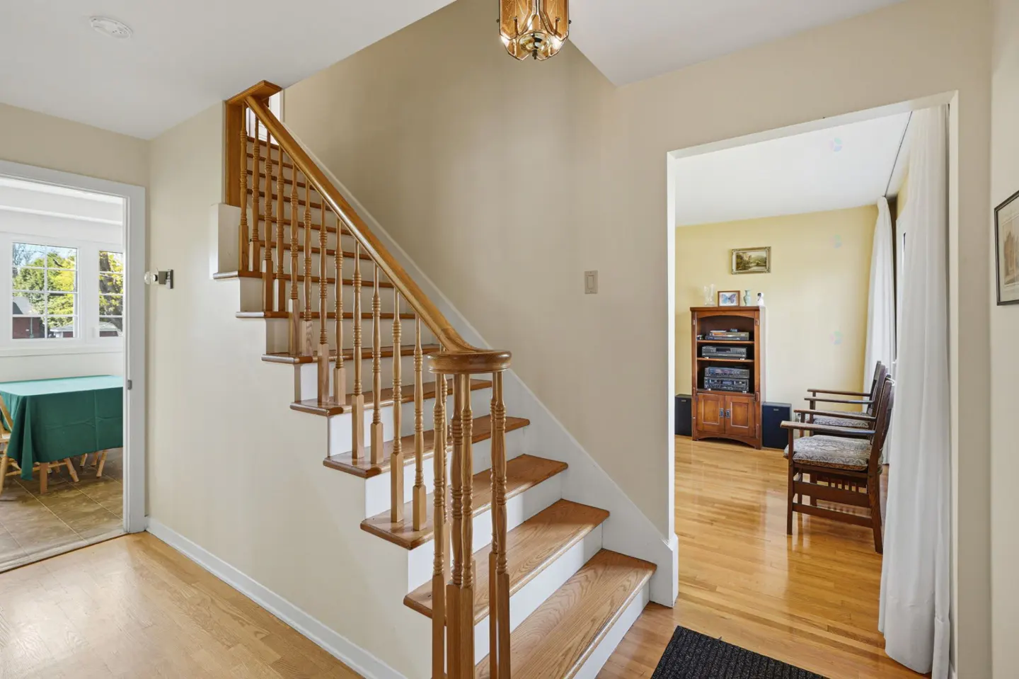 A bright foyer with a wooden staircase, light beige walls, and hardwood floors. A dining room and living room are visible through doorways.