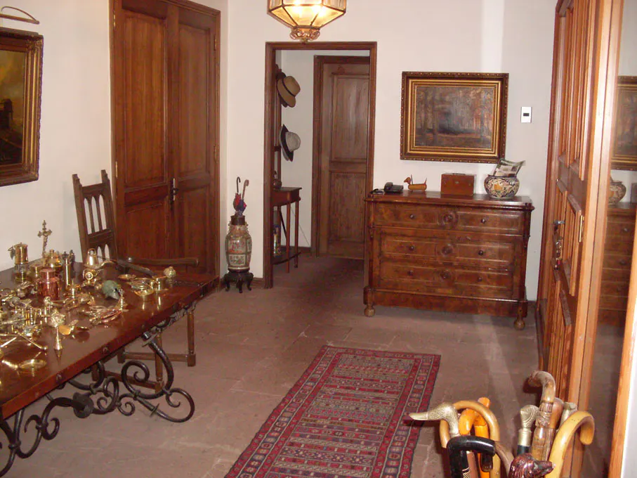 A room with antique furniture, including a wooden table covered in brass objects, a chest of drawers, and a rug on a terracotta floor.