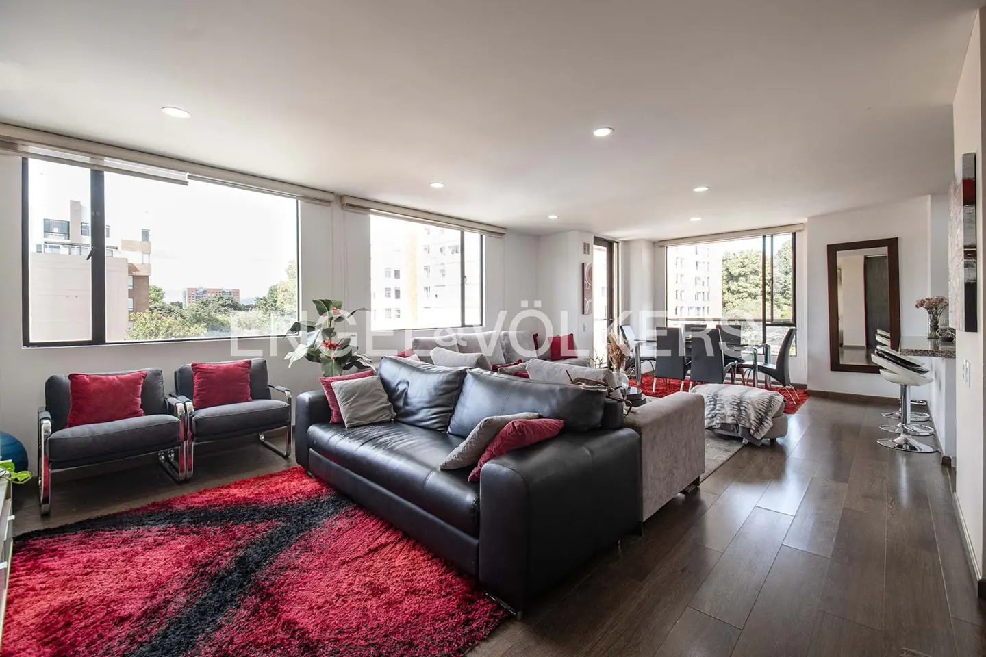 Bright living room with a black leather sofa, red rug, and large windows overlooking a cityscape.