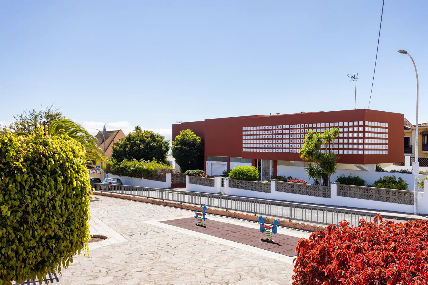 Exterior view of a modern, two-story house with a red facade and white square accents, overlooking a playground with spring riders.